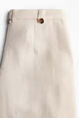 Pantalón sastrero cropped de tiro alto, color beige, con piernas amplias, pliegues frontales, bolsillos al bies y trabillas en la cintura con detalles de botones.
