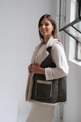 Chaleco de gamuza sintética en color verde oliva con interior y bordes de corderito beige. Presenta solapas amplias, dos bolsillos frontales aplicados y un diseño abierto sin cierre.
