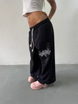 Pantalón de jogging negro de corte baggy, confeccionado en algodón. Presenta una estampa plastisol con bordado decorativo en la pierna izquierda, cintura elástica con cordón largo ajustable y botones en el ruedo para regular la botamanga.