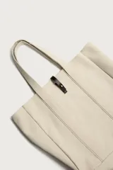 Bolso tipo shopper de tejido color crudo, con doble asa de hombro y detalles en cuero negro. Cuenta con compartimento principal con cierre de tira y botón a presión.