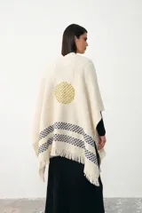 Ruana de tejido de punto grueso en color crudo, con diseño de círculo bordado en tono mostaza en la parte central superior y franjas horizontales con patrón geométrico en azul marino en la parte inferior. Presenta terminaciones con flecos en el ruedo.