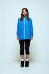 Camisa azul de corte oversize, con cierre frontal de botones y detalles de cintas onduladas en colores contrastantes a lo largo de la botonadura y en los puños. Presenta cuello clásico y mangas largas.