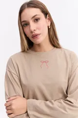 Remera de manga larga color beige con silueta relajada y mangas amplias. Presenta una estampa en la espalda con la frase 'Be kind to your mind' acompañada de ilustraciones de un cupido, flores y lazos en tono bordó.