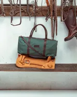 Bolso tipo duffel de gran tamaño, confeccionado en lona color verde oliva con detalles y correas en cuero color suela. Cuenta con cierre superior y etiqueta de cuero con el logo de la marca.