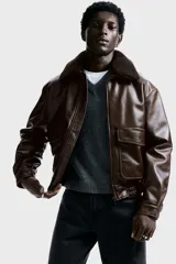 Campera de estilo aviador confeccionada en tejido revestido con acabado de cuero sintético. Presenta cuello de sherpa en contraste, cierre frontal de cremallera, hombros caídos y bolsillos delanteros de parche con solapa y botón a presión. Incluye elástico fruncido en el bajo y puños ajustables con botón a presión.