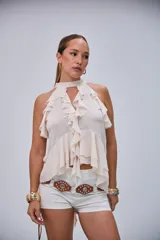 Blusa sin mangas de color crudo con cuello halter y abundantes volados en la parte frontal y sisas.