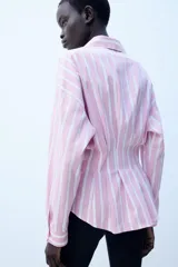 Camisa de popelina con diseño a rayas verticales rosadas y blancas, cuello solapa con escote en V, manga larga y detalle de pliegues marcados en la cintura. Cierre frontal con botones ocultos.