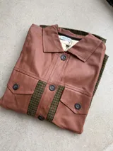Leñadora de paño con patrón de espiga en tonos verde y marrón, combinada con detalles de cuerina habano en el canesú frontal, solapas, cuello y bolsillos plaqué con solapa. Presenta cierre frontal de botones oscuros y mangas largas con puños abotonados.