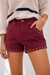 Short de tiro medio con efecto gamuza en color bordó, decorado con tachas metálicas doradas en los bordes y bolsillos. Cuenta con presillas para cinturón y cierre frontal.