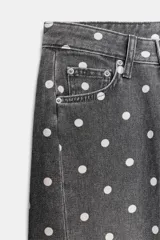 Jeans de tiro medio con corte relajado y largo al tobillo. Presenta un diseño de cinco bolsillos, cierre frontal con cremallera y botón metálico, y un estampado integral de lunares blancos sobre fondo gris oscuro.