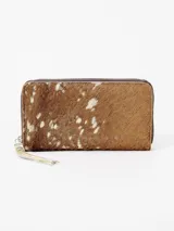 Billetera grande de cuero con pelo, con estampado animal print tipo vaca en tonos marrones y blancos. Cuenta con cierre perimetral y tirador de cuero con flecos.