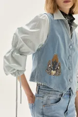 Chaleco de denim celeste con bordados decorativos en el frente, cierre de botones y corte entallado.