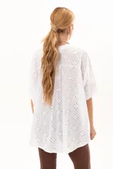 Camisa de broderie blanca con silueta oversize, escote en V, cierre frontal con botones y mangas 3/4. Presenta un diseño con bajo asimétrico, siendo ligeramente más larga en la parte posterior.
