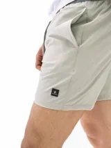 Short de baño de secado rápido, color verde claro o gris pálido, con cintura elástica y cordón de ajuste verde. Presenta un pequeño parche negro con un logo en la parte inferior derecha del frente.