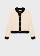 Chaqueta de punto de textura suave en color crema con detalles en contraste negro en cuello, puños, ruedo y tapeta de botones. Presenta un diseño recto, cuello redondo y cierre frontal con botones dorados.