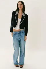 Blazer corta de color negro con cuello solapa, manga larga y hombreras. Presenta detalles combinados con estampado de cuadro vichy en el cuello, los bordes de la solapa, el ruedo y los puños. Cuenta con cierre frontal de un botón y falsos bolsillos delanteros de vivo.
