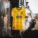 Camiseta de fútbol Puma del Club Peñarol, modelo Away Jersey. Diseño amarillo con detalles en negro en el cuello en V, los bordes de las mangas y líneas horizontales en el pecho. Incluye el logo de Puma, el escudo del club y el sponsor Antel.