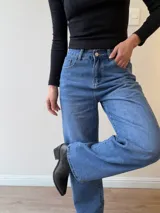 Pantalón de jean corte mom fit con tiro alto, confeccionado en denim de lavado clásico. Presenta cierre frontal con botón y cierre metálico, con bolsillos funcionales delanteros y traseros.