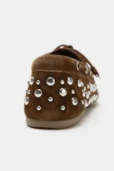 Zapato tipo náutico de serraje color marrón, con detalle de tachas metálicas plateadas en todo el cuerpo y cordones decorativos en la parte delantera. Presenta punta redonda y suela plana.
