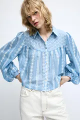 Blusa corta de color blanco con diseño de bordados perforados, cuello solapa, cierre frontal con botones y mangas largas abullonadas.
