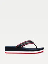 Ojotas de mujer marca Tommy Hilfiger con suela de cuña. Cuentan con tiras de nailon trenzado en color azul marino con detalles en rojo y blanco, firma de la marca bordada y logo metálico. La suela de goma presenta franjas decorativas en los colores distintivos de la marca.