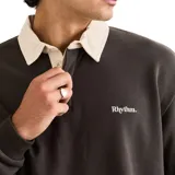 Buzo tipo polo de rugby con mangas largas, confeccionado en algodón color gris oscuro. Presenta cuello de sarga en color crema en contraste, cierre frontal parcial con botones y logo bordado pequeño en el pecho.