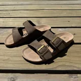 Sandalia tipo Birkenstock con doble tira ajustable de cuero sintético negro y hebillas metálicas. Cuenta con plantilla anatómica de corcho y suela de goma.