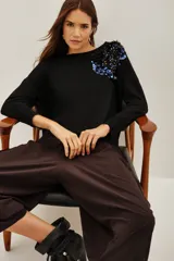 Sweater negro de punto con cuello redondo y mangas largas. Presenta una aplicación decorativa de lentejuelas y pedrería en tonos azules y oscuros sobre el hombro izquierdo.