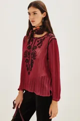 Camisa de manga larga en color negro con tejido semitransparente y detalles de hilos metalizados. Presenta un diseño con bordados florales en tonos tierra, cierre frontal con botones y un lazo decorativo en el cuello.