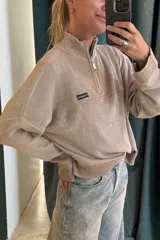 Sweater de tejido acanalado color beige, con cuello alto y cierre parcial frontal. Presenta un pequeño parche negro con texto blanco en el pecho.