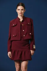 Saco corto color bordó con cuello mao, cierre frontal de botones dorados y cuatro bolsillos delanteros con solapa y botón dorado. Presenta un parche decorativo en la manga izquierda.