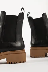 Bota tipo chelsea con suela track de goma en color marrón y capellada de cuero negro. Presenta elásticos laterales y tirador trasero.