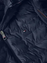 Chaleco azul marino acolchado con capucha, cierre de cremallera frontal y dos bolsillos laterales con cremallera. Tiene el logo de Tommy Hilfiger bordado en el pecho y detalles distintivos de la marca.