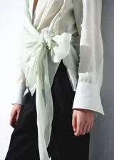 Camisa cruzada de ramio en color crudo, con cuello camisero, escote en V y mangas largas con puños anchos abotonados. Cuenta con un diseño envolvente con detalle de lazo lateral.