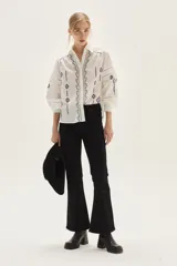 Blusa blanca de algodón con bordados geométricos en tonos negros y lilas, cuello clásico, cierre frontal con botones y mangas largas abullonadas con puños. Presenta detalles de vainillas y calados en el frente y mangas.