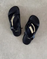 Sandalias planas de color negro con tiras finas y detalle decorativo de tubos metálicos dorados en el empeine. Cuentan con cierre de hebilla ajustable en el tobillo.