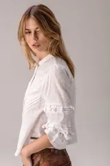 Blusa blanca de broderie con cuello alto con volados, mangas largas abullonadas con detalles de encaje y pliegues decorativos en el frente.