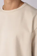Remera de corte oversize color beige, con cuello redondo y etiqueta decorativa pequeña en el ruedo inferior.