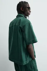 Camisa de denim de algodón color verde oscuro, de corte relajado (relaxed fit) y manga corta. Posee cuello solapa, bolsillo plastrón en el pecho con bordado a tono y cierre frontal de botones.
