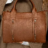 Cartera de cuero color tostado, estructurada, con doble asa de mano, correa desmontable y dos bolsillos frontales verticales con cierre. Presenta detalles de costuras pespunteadas y el logo de la marca grabado en la parte inferior.