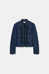 Blazer corto de tweed en color azul, con cuello mao y manga larga. Presenta detalles de pasamanería negra en el frente y cierre con ganchos metálicos y falsos botones dorados.