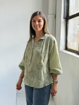 Camisa verde claro de corte oversize, con mangas tres cuartos abullonadas, escote en V con cuello camisero y cierre frontal parcial de botones. Cuenta con delicados bordados florales tono sobre tono distribuidos por toda la prenda.