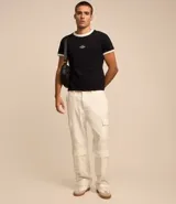 Remera blanca de algodón, modelo baby tee, con mangas cortas, cuello redondo y detalles contrastantes en marrón. Presenta un pequeño logo gomoso con la palabra "Lucky" en el centro del pecho.