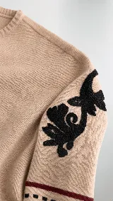 Sweater de lana color beige con calce holgado y cuello redondo. Presenta un bordado negro con diseño de arabescos en una de las mangas y detalles de rayas y puntos en contraste en el ruedo y los puños.