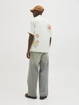 Camisa de algodón color crudo con estampado de flores y billetes, cuello resort, calce relajado, mangas cortas y cierre de botones.