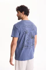 Remera azul de manga corta con estampa en la espalda de diseño tipo estampilla con paisaje de palmeras, olas y montañas.