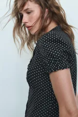 Camisa negra con estampado de lunares blancos, cuello redondo con escote en pico, mangas cortas abullonadas y detalle de volantes frontales. Cierre frontal con botones.