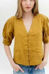 Blusa de lino color mostaza con escote en V y cierre frontal de botones forrados. Presenta mangas abullonadas al codo con puños anchos y detalles de costuras verticales en el frente.
