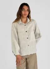 Chaqueta color crema de lino con cuello camisero, cierre frontal con botones, mangas largas con puños abotonados, bolsillos de parche en el pecho y bajo con flecos.