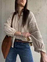 Camisa beige de inspiración boho con detalles bordados en mangas y puños, cuello en V con botones frontales y mangas largas abullonadas.